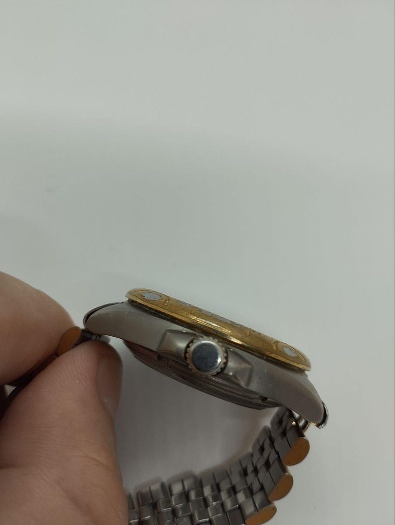 May include: A silver and gold wristwatch with a silver band. The watch face is silver with a gold bezel. The watch is being held by a person's hand.