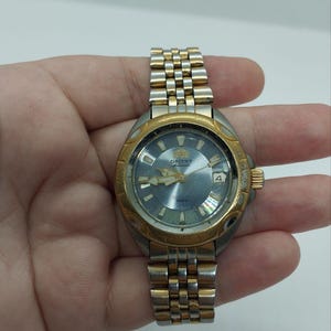 May include: A vintage Orient watch with a gold-tone bezel and a silver and gold-tone band. The watch face is a light blue color with gold-tone hands and markers. The watch has a date display at the 3 o'clock position.