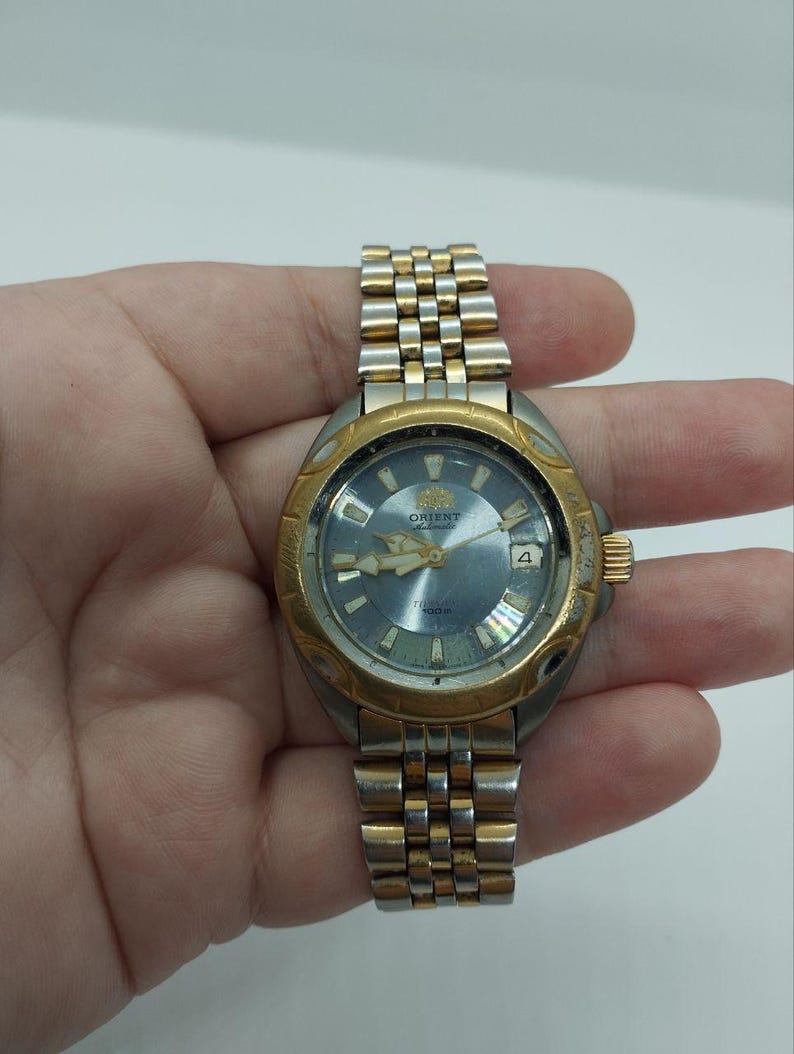 May include: A vintage Orient watch with a gold-tone bezel and a silver and gold-tone band. The watch face is a light blue color with gold-tone hands and markers. The watch has a date display at the 3 o'clock position.