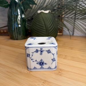 May include: A square, white ceramic bud vase with blue floral patterns. The vase has a small opening at the top and is decorated with a delicate blue floral design. The vase is sitting on a wooden surface.