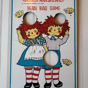Può includere: Gioco vintage del lancio dei sacchi di fagioli di Raggedy Ann e Andy. Il tabellone presenta illustrazioni dei personaggi, tre fori bersaglio e valori dei punti. Il tabellone ha un bordo blu e il testo "Raggedy Ann and Andy Bean Bag Game".