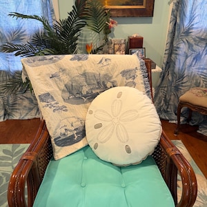 May include: A wicker armchair with a turquoise cushion and a round, white sand dollar pillow. A cream-colored throw blanket with blue nautical illustrations is draped over the chair. The scene is set in a room with tropical decor.