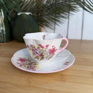 May include: A white porcelain teacup and saucer set with a scalloped edge and a pink handle. The set features a delicate floral pattern with clusters of purple, pink, and white flowers. A thin pink line accents the rim of both the cup and the saucer.