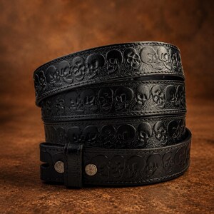 Op de afbeelding: Een zwarte leren riem met een herhalend reliëfpatroon van schedels over de gehele lengte van de band. De riem is voorzien van een bijpassende leren lus en twee zilverkleurige metalen klinknagels die het uiteinde van de band vastzetten.