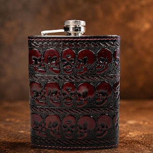 May include: A rectangular metal flask wrapped in dark leather featuring an embossed pattern of multiple skulls arranged in horizontal rows. The leather is dyed in deep red and black tones with visible stitching and a silver-coloured screw cap.