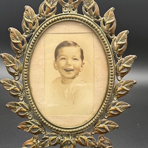 Könnte beinhalten: Ein antiker ovaler Bilderrahmen mit einem Vintage-Schwarz-Weiß-Foto. Der Rahmen besteht aus goldfarbenem Metall mit einem Blattdesign um die ovale Öffnung. Das Foto zeigt ein lächelndes Kind.
