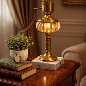 May include: A table lamp featuring a brass base with a twisted column, a ribbed amber glass shade, and a perforated brass top detail, all mounted on a square white marble base. The lamp emits a warm glow from an internal light source.