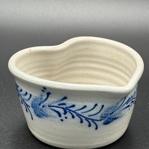 Puede incluir: Un pequeño cuenco de cerámica blanquecina con borde en forma de corazón. El exterior presenta un patrón floral azul alrededor de la base. El interior no está esmaltado y el cuenco tiene un aspecto rústico y hecho a mano.