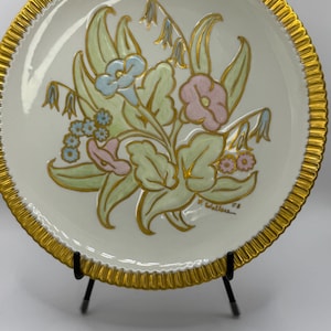 May include: Cream-colored decorative plate with a gold rim and floral design. The flowers are in shades of pink, blue, and green, with gold accents. The plate is on a black metal stand.