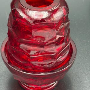 Pode incluir: Um castiçal de vidro vermelho rubi com um design geométrico. O castiçal tem uma base larga e um topo cilíndrico, com uma superfície texturizada e facetada. O vidro é transparente, permitindo a passagem da luz.