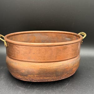 May include: A round copper basin featuring a wide, smooth rim and a slightly rounded body. The vessel is equipped with two curved brass handles attached to opposite sides. The copper surface displays a natural, weathered patina with varied tones.