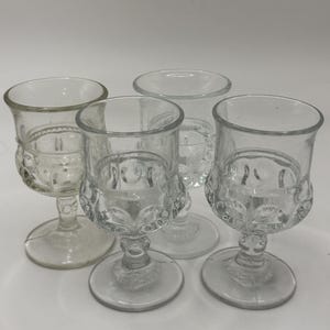 Puede incluir: Colección de cinco copas de cristal transparente. Cada copa presenta una base redonda, un tallo con un diseño decorativo y un borde acampanado. Las copas están dispuestas sobre una superficie blanca, mostrando su intrincado diseño y transparencia.