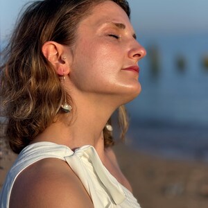 May include: A gold-toned drop earring featuring a delicate chain that suspends a small, white spherical pearl nestled within a dark, textured shell-like base. The earring is worn in the earlobe, showcasing a slender metal wire design.