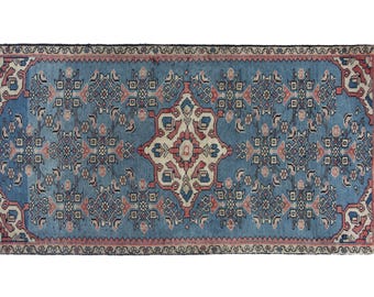 Puede incluir: Alfombra rectangular azul con un patrón intrincado. La alfombra presenta un diseño de medallón central en crema y rojo, rodeado de motivos florales y geométricos repetidos en tonos de azul, rosa y crema. El borde es azul oscuro con un ribete rosa.