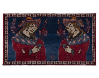 Puede incluir: Alfombra rectangular con un campo central azul y un borde. La alfombra presenta un diseño simétrico de dos figuras con túnicas rojas, sosteniendo tazas. Las figuras tienen pelo largo y llevan sombreros. El borde es azul oscuro con patrones florales y geométricos.