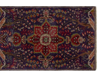 Puede incluir: Una alfombra rectangular con un fondo morado intenso y un medallón floral central en rojo y crema. El diseño incluye intrincados patrones botánicos en tonos verde, naranja y azul claro, rematado con flecos cortos en los extremos.