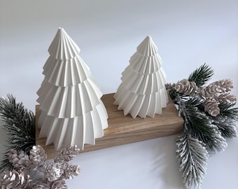 May include: Two white, cone-shaped, pleated tree ornaments of varying heights mounted on a rectangular wooden base. The display is accented with artificial evergreen branches featuring frosted needles and small, light-colored pine cones.