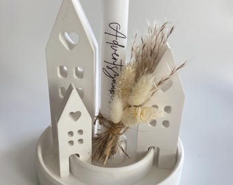 May include: A white Advent candle with the text "Adventsgruss" and decorative white ceramic houses with heart-shaped cutouts. The candle is surrounded by dried flowers and sits on a white, circular base.