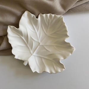 Könnte beinhalten: Eine weiße, blattförmige Keramikschale mit detaillierten Adern. Die Schale ist vor einem neutralfarbenen Stoffhintergrund platziert, was eine schlichte und elegante Ästhetik erzeugt. Diese dekorative Schale ist ideal zum Servieren kleiner Snacks oder als dekoratives Element.