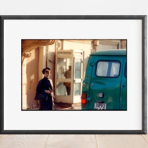 May include: A woman in a black coat and hat walks past a green van with a license plate that reads "3892".