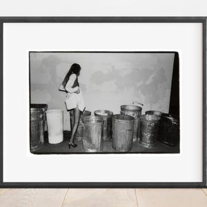 May include: A monochrome framed photograph showing a person standing beside a collection of metal bins. The image is a medium shot, with the person centrally positioned and the bins in the foreground. The photograph is presented in a black frame.