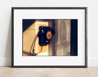 May include: A framed photograph of a vintage black rotary dial telephone mounted on a wall. The phone has a circular dial and a wooden center. The image is lit by sunlight, casting shadows on the wall and the phone's cord.