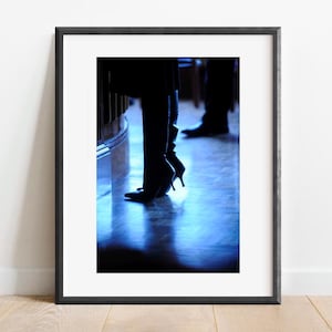 May include: A pair of black heeled boots with a pointed toe, one boot is slightly higher than the other, standing on a wooden floor with a blue light reflecting off the surface.