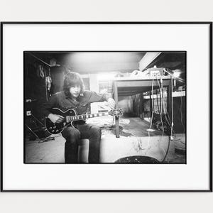 Könnte beinhalten: Schwarz-weiß gerahmtes Foto einer Person, die eine E-Gitarre spielt. Die Person sitzt, und die Gitarre liegt auf ihrem Schoß. Der Hintergrund umfasst einen Tisch mit Ausrüstung und Kabeln.