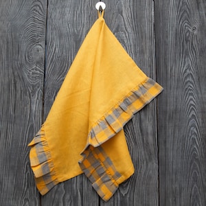 May include: A yellow linen kitchen towel with gray plaid trim and ruffled edges hangs from a silver hook against a dark wood background. The towel is folded, showcasing the contrasting colors and textures.