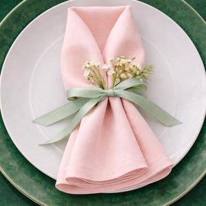 Può includere: Un tovagliolo di lino rosa piegato, adornato con un rametto di fiori bianchi e un nastro verde salvia, è appoggiato su un piatto bianco. Il piatto è su un sottopiatto verde, creando un'elegante mise en place. Ideale per eventi.