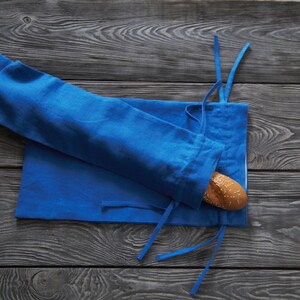 May include: A blue linen bread bag with a baguette inside. The bag is tied with a blue ribbon and is sitting on a wooden surface.