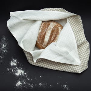 May include: A brown loaf of bread wrapped in a white and beige checkered linen cloth. The cloth is folded over the bread, and the edges of the cloth are visible around the bread.