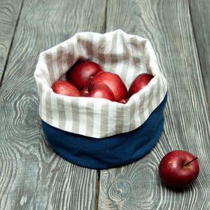 May include: A navy blue fabric basket with a striped interior holds several red apples. One apple rests outside the basket on a gray wooden surface. The basket's interior lining features vertical stripes in beige and white.