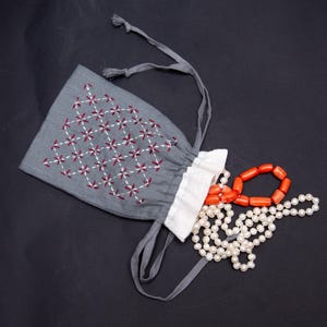 May include: A gray drawstring pouch featuring a white and maroon embroidered geometric floral pattern. The pouch has a white ruffled top edge and two gray fabric ties. A strand of white pearls and a strand of orange coral beads spill out.