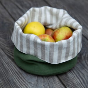 May include: A green fabric basket with a striped linen lining, filled with yellow and red apples. The basket sits on a dark wooden surface, creating a rustic and natural aesthetic. The apples are ripe and fresh.