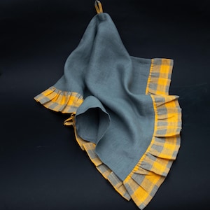 May include: A grey linen kitchen towel with a yellow and grey plaid ruffle trim. The towel has a loop for hanging and is draped against a dark background. The ruffle trim adds a decorative element to the towel.