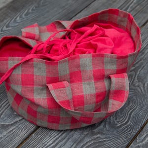 Puede incluir: Una bolsa de lona de cuadros rojos y grises con forro rojo. La bolsa está doblada y colocada sobre una superficie de madera.