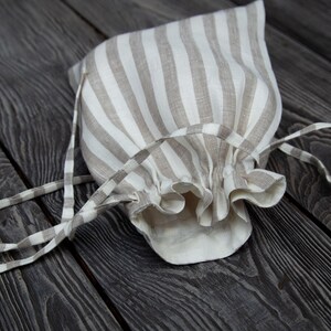 May include: A beige and white striped linen drawstring bag with a gathered top. The bag is lying on a dark wood surface.