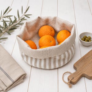 Op de afbeelding: Een ronde stoffen mand met een beige en wit verticaal gestreepte buitenkant en een effen beige linnenachtige voering. De mand is gevuld met vijf feloranje citrusvruchten en heeft een zachte, getextureerde textielconstructie.