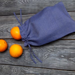 May include: A blue drawstring bag with two oranges partially visible and two oranges beside it. The bag is made of a soft fabric and is partially open, with one orange peeking out. The oranges are bright orange and have a smooth texture.