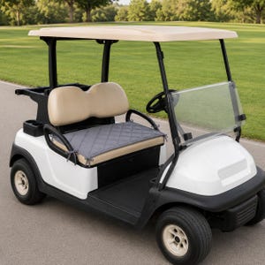 May include: A white golf cart featuring a beige roof, beige seats, and a black steering wheel. A grey quilted seat cushion with tie-fastenings is placed on the bench seat. The vehicle has black trim, black wheels, and a clear windshield.