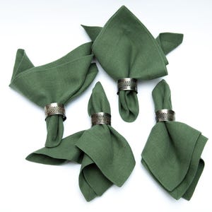 Puede incluir: Cinco servilletas de lino verde con anillos de servilleta plateados. Las servilletas están dobladas de diferentes maneras.