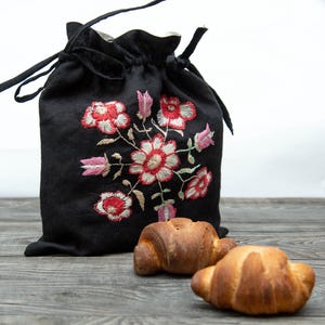 May include: A black drawstring bag with embroidered red and white flowers and pink accents. Two golden-brown croissants rest in front of the bag on a gray wooden surface. The bag is made of a soft, dark material.