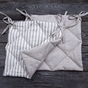 May include: Two beige linen chair cushions with one side featuring a white and beige striped pattern and the other side featuring a quilted design. The cushions have ties at each corner.