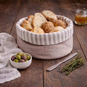 May include: A linen bread basket with a striped lining, containing assorted breads. A small bowl of olives, a knife, and thyme sprigs are on a wooden surface, with a jar of preserves in the background.