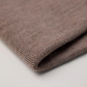 Puede incluir: Una pieza doblada de tela de punto de color marrón claro con una textura fina y consistente. El material presenta una apariencia jaspeada sutil con nervaduras verticales visibles a lo largo del borde, mostrando una construcción tejida.