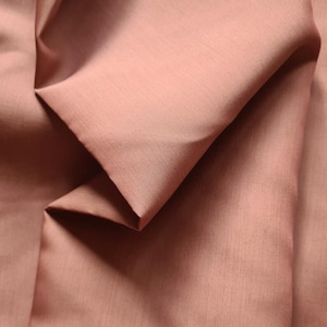 Peut inclure: Un tissu rose poudré uni avec une texture lisse et mate. Le matériau est drapé en plis naturels et doux, créant des ombres et des reflets subtils sur la surface qui soulignent la couleur rose brunâtre, sourde et uniforme du textile.