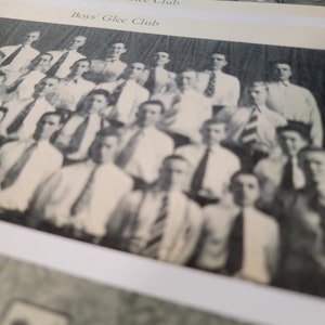 Op de afbeelding: Zwart-witfoto van een middelbare school zangkoor. De foto is verdeeld in twee secties, een voor het meisjeskoor en een voor het jongenskoor. Het meisjeskoor staat in een rij, terwijl het jongenskoor in een halve cirkel staat.