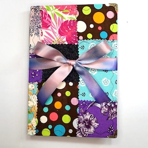 May include: A fabric-covered journal with a patchwork design featuring floral, polka dot, and patterned squares. A satin ribbon in shades of pink and purple is tied around the journal. The journal's corners have gold-colored metal protectors.