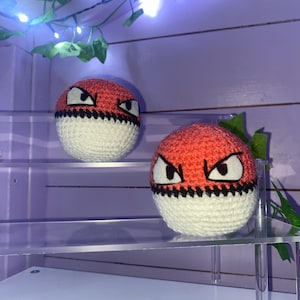 Può includere: Due Poké Ball di lana rossa e bianca con occhi neri. Le Poké Ball sono appoggiate su uno scaffale in acrilico trasparente.